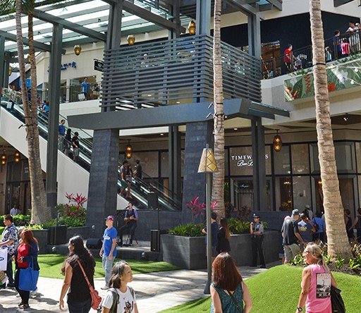 People walking around outdoors at Ala Moana shopping center on a sunny day. Bright green grass in the foreground. An escalator to second floor shows in the background.